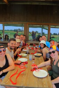 Groupe de personnes souriantes assises autour d'une table en bois pour un repas en plein air avec vue sur un paysage champêtre.