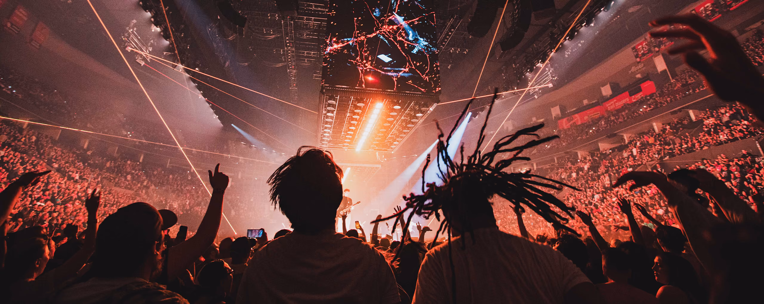 An energized crowd in an arena silhouetted against lights and lasers with Linkin Park performing on stage 