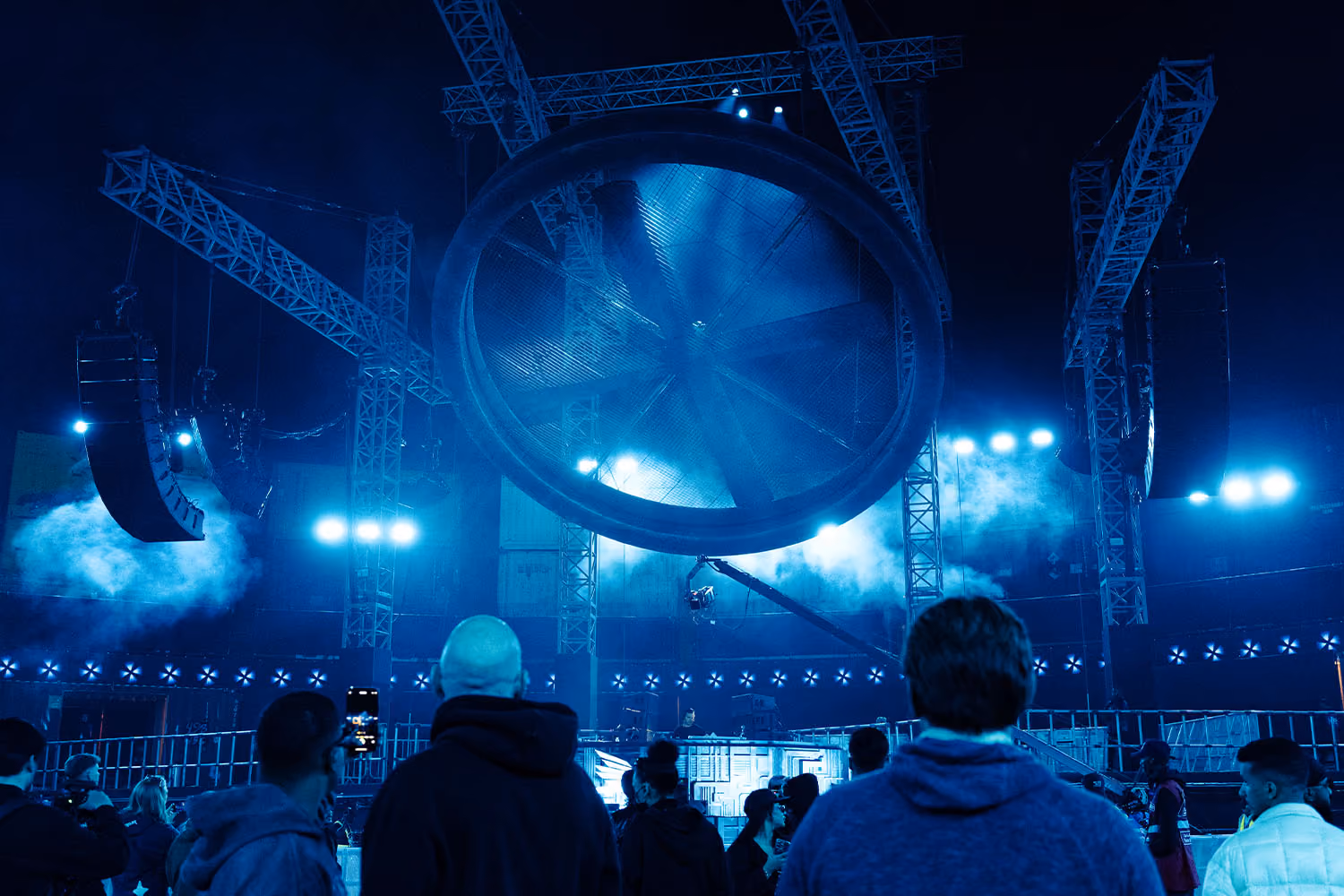 A large crowd gathers under a massive wheel set piece at MDLBeast Soundstorm, with a large Cohesion PA system suspended overhead