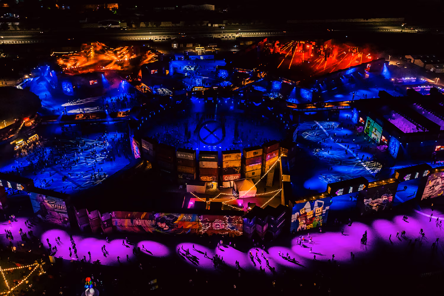 Aerial view of MDLBeast Soundstorm's Underground stages, illuminated by a striking display of purple and blue lights. The festival grounds are enclosed by metal shipping container walls adorned with graffiti and artwork