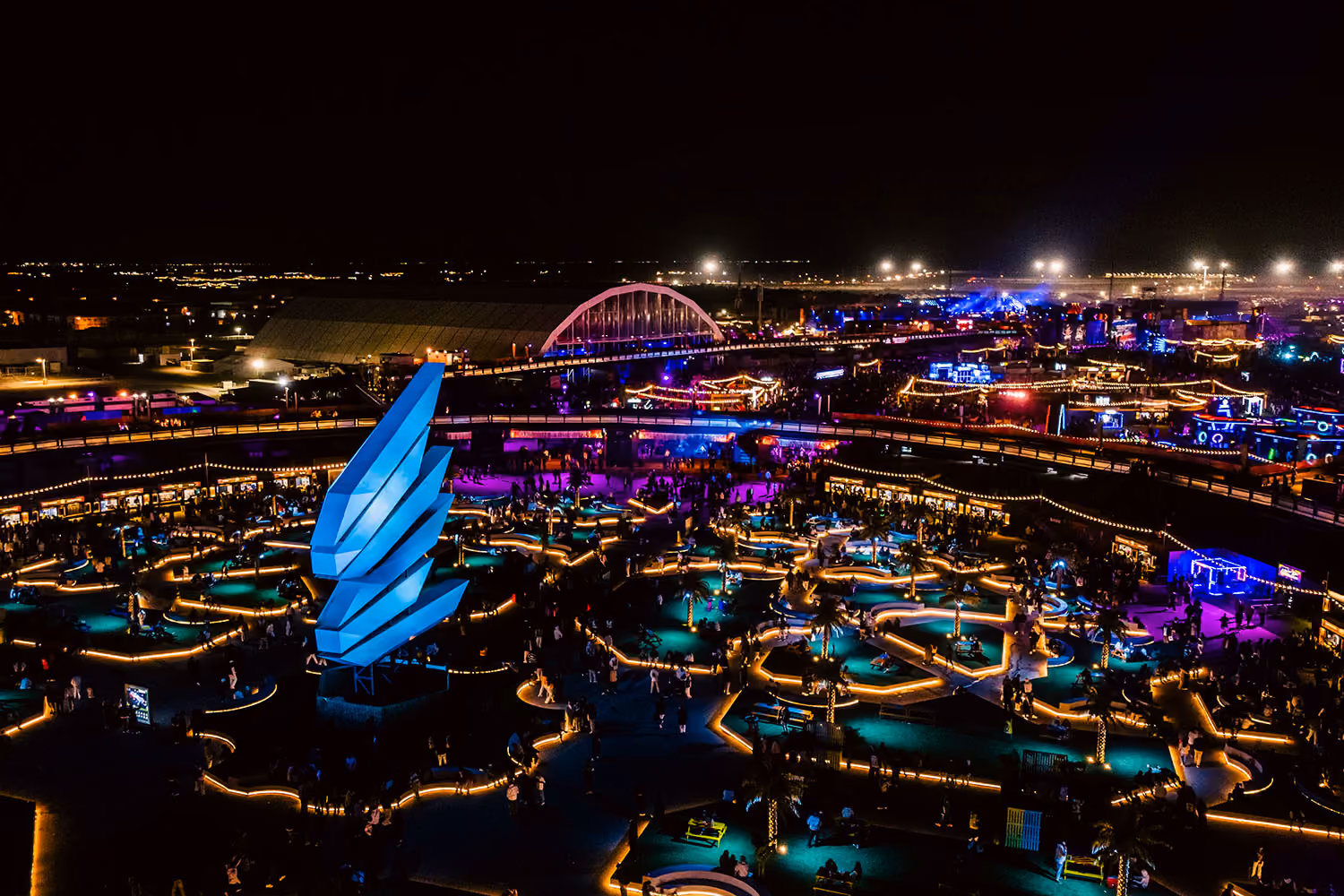 Aerial night view of the MDLBeast Soundstorm festival grounds, glowing with vibrant lights and featuring a massive illuminated MDLBeast logo in blue.