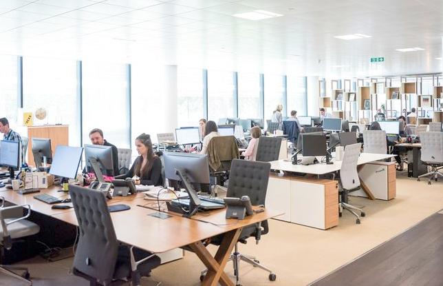 Großraumbüro mit zahlreichen Schreibtischen, Monitoren, Bürostühlen und fleißigen Mitarbeitern