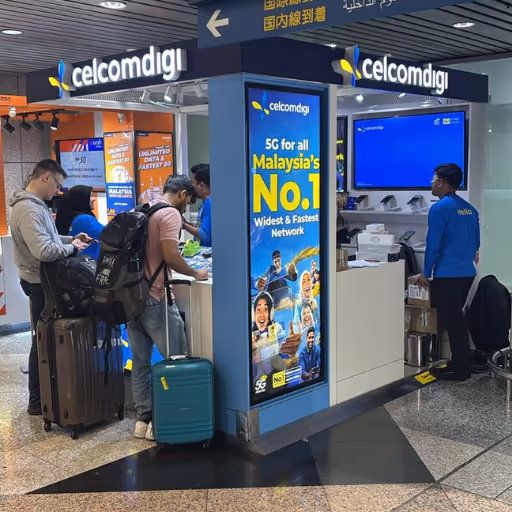 CelcomDigi outlet at KLIA Terminal 1, showing travelers being assisted at the counter with promotional signage for Malaysia’s No. 1 5G network and staff members wearing blue uniforms.