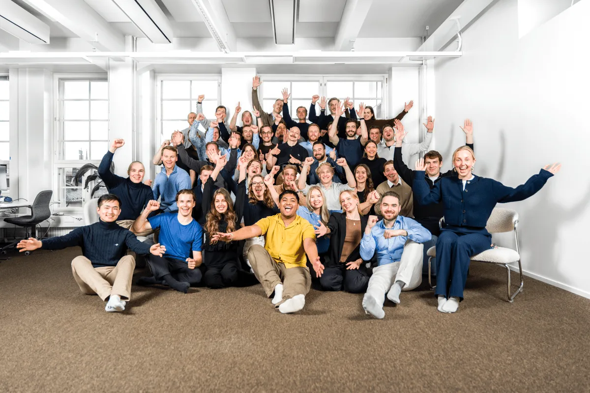 Group photo of the entire company team, with over 40 people smiling and raising their hands in celebration. Taken in a modern office with large windows, the team is arranged in multiple rows, creating a dynamic and energetic atmosphere.

