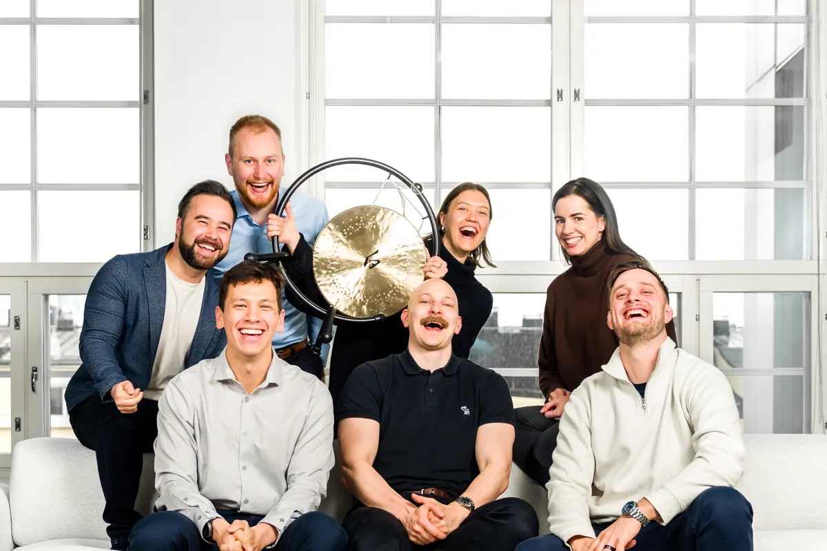 Photo of the commercial team featuring seven people in a cheerful pose, gathered around a golden gong. Everyone is laughing and smiling in a bright, modern office, creating a fun and energetic vibe.
