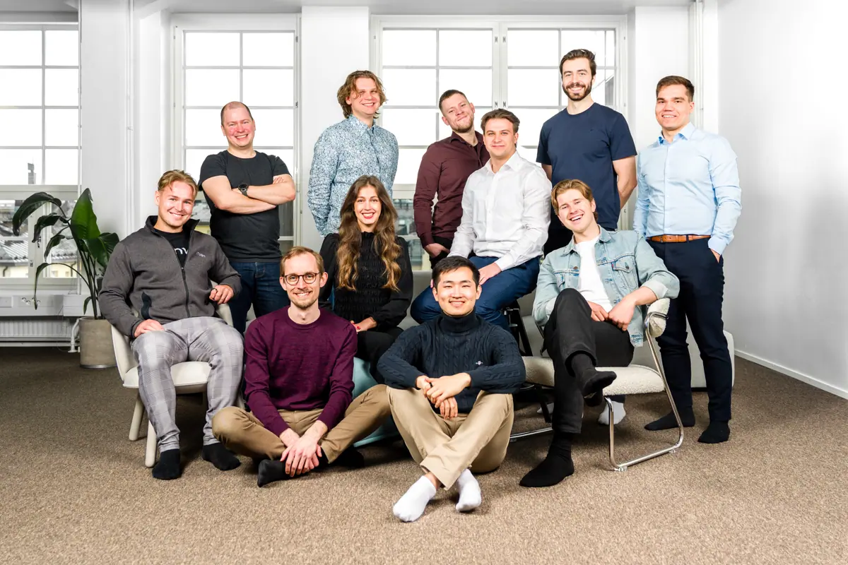 Group photo of the development team with twelve members posing casually in a well-lit office. The team is a mix of seated and standing people, dressed in relaxed business-casual clothing and smiling confidently.
