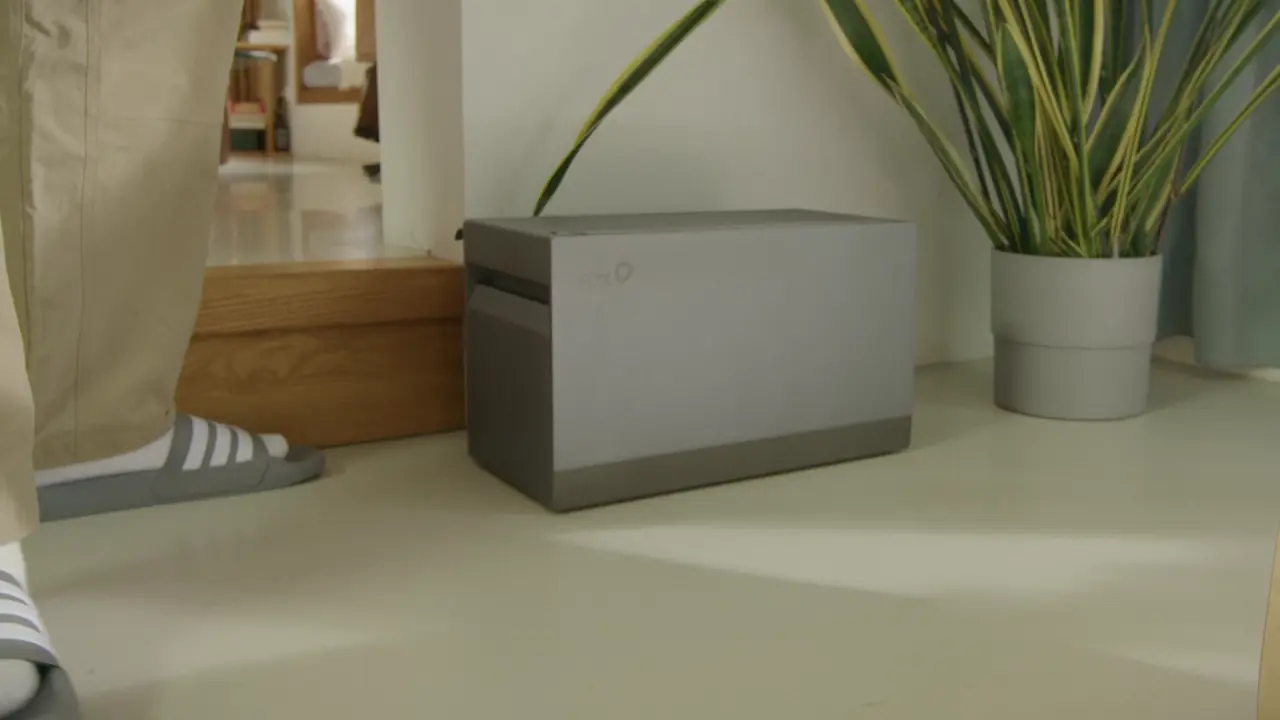 Een grijze thuisbatterij met stekker van het merk FoxESS staat in een woonkamer naast een plant. Naast de batterij staat een persoon in een beige broek, witte sokken en grijze slippers.