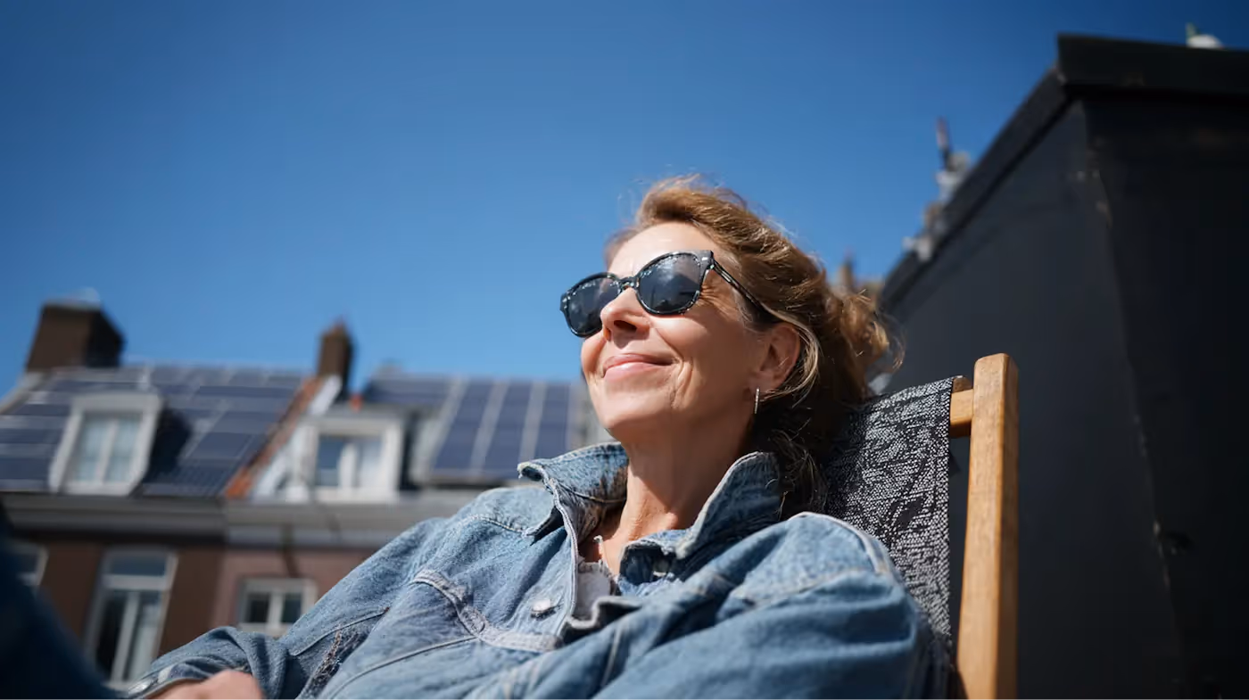 Een vrouw met zonnebril zit op een dakterras in het zonlicht.