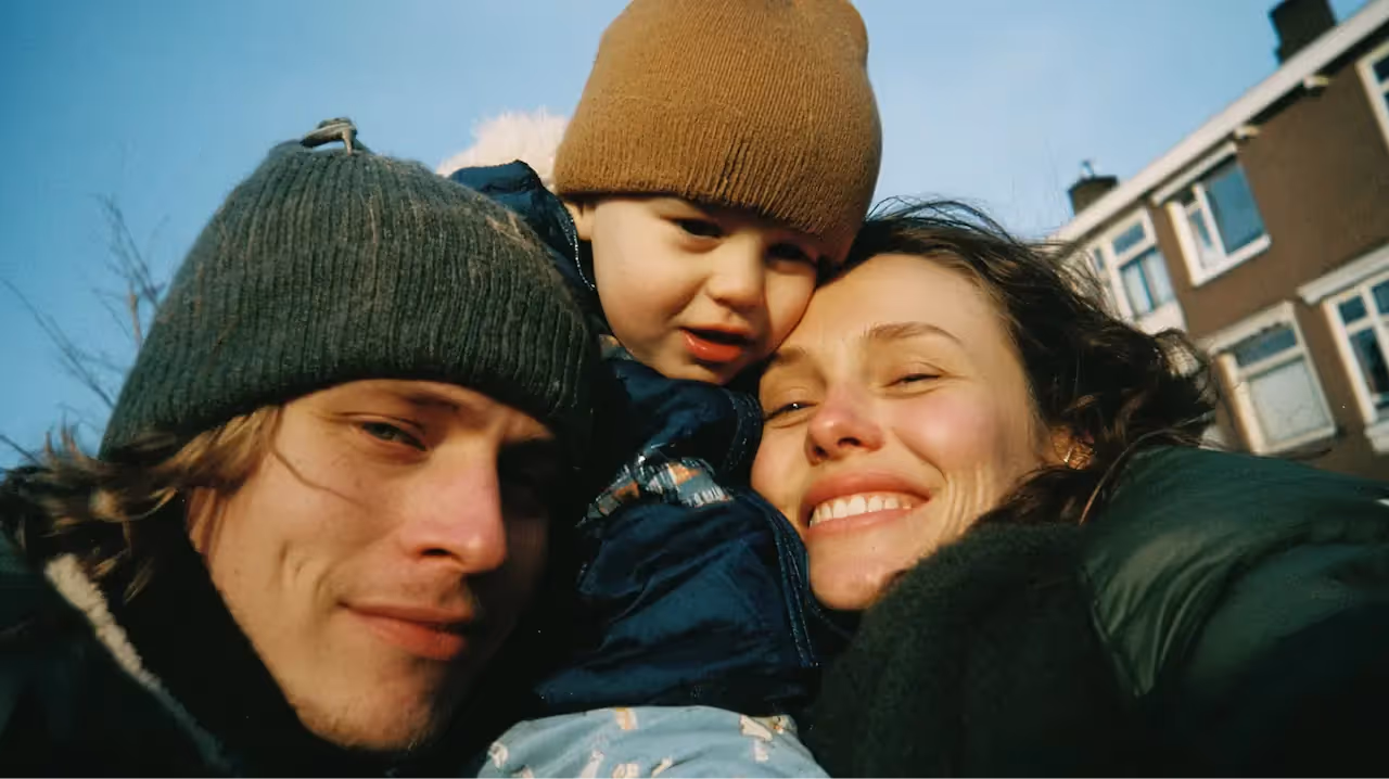 Twee ouders poseren voor een selfie met hun kind, alledrie gekleed in winter kleding.