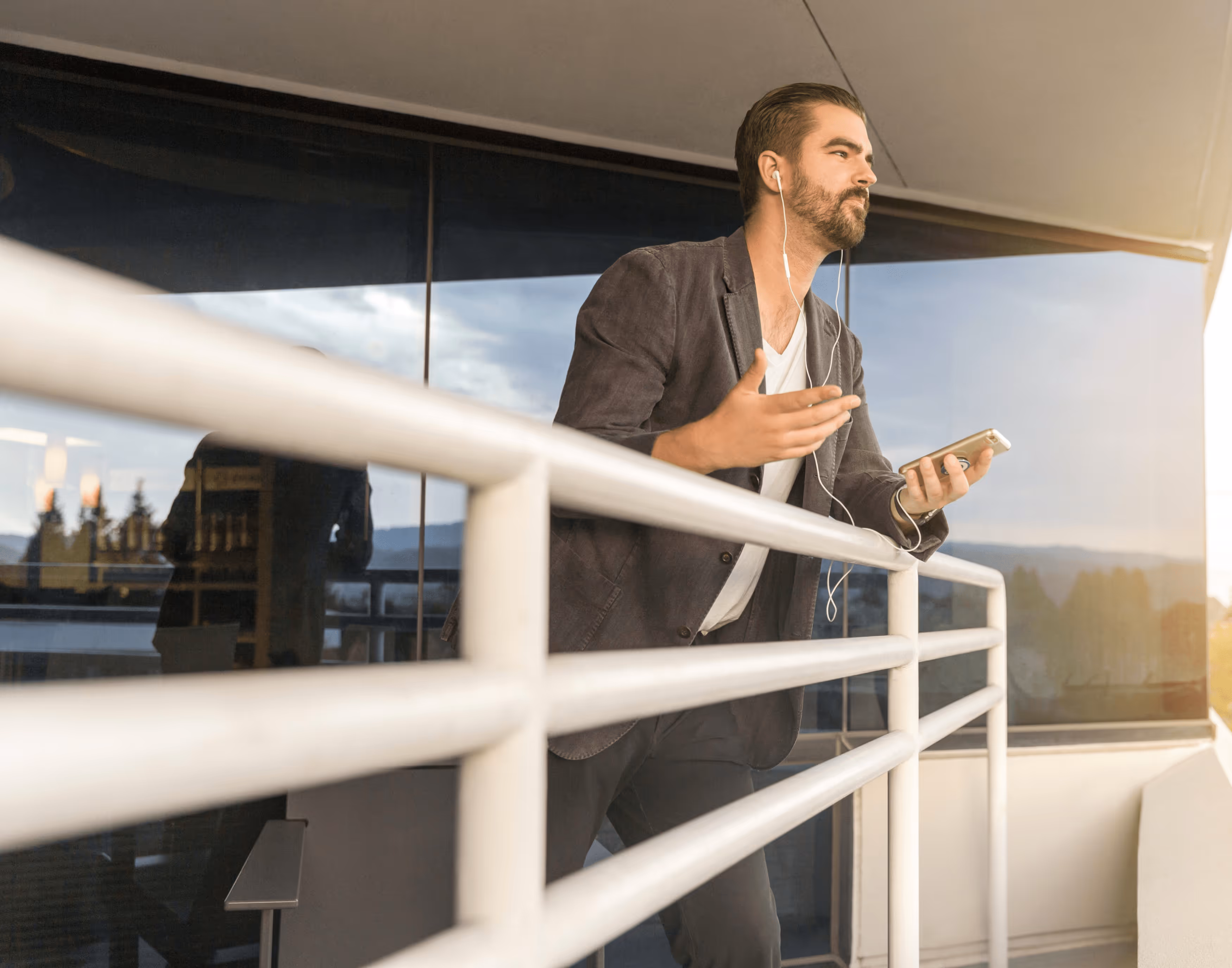 Man wearing a blazer and earphones speaking while holding a smartphone on a balcony with glass windows in the background.