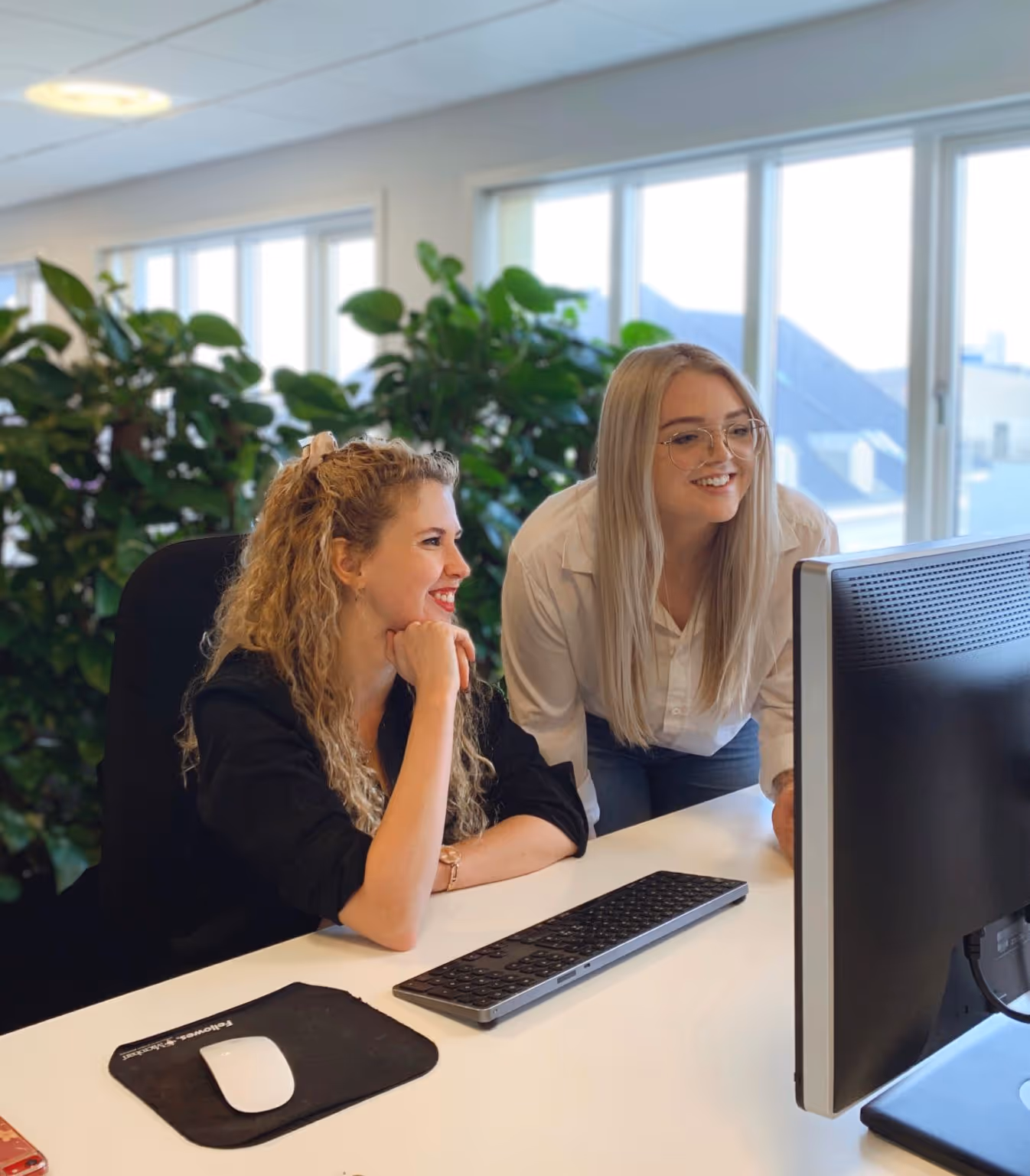 Two smiling women looking at a computer screen in a bright office with large windows and green plants.