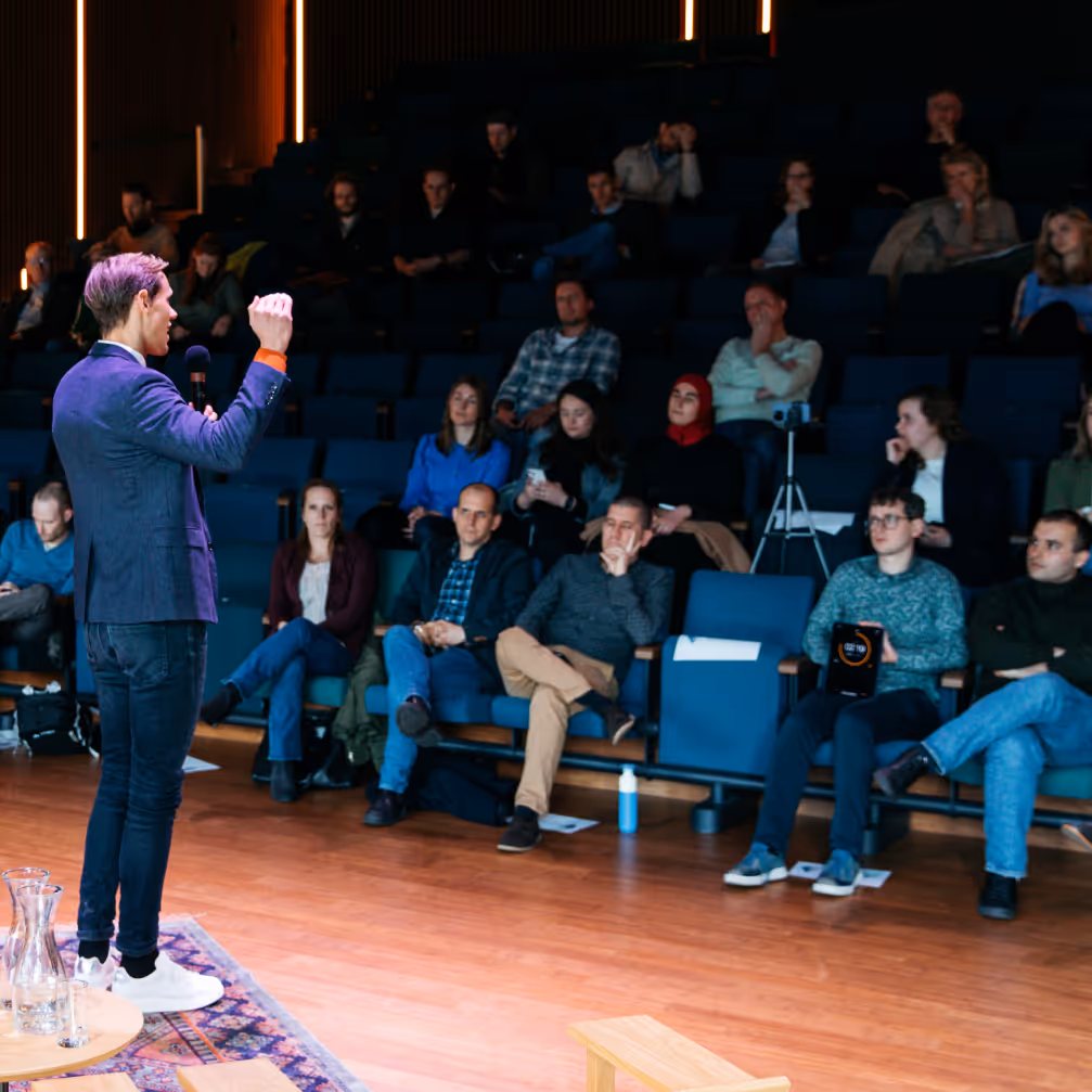 Speaker in casual attire holding a microphone and addressing an attentive seated audience in an auditorium.