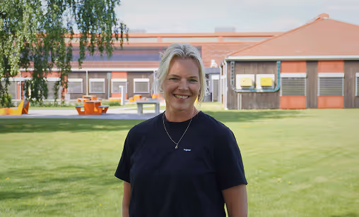 Smiling woman with blonde hair wearing a black shirt standing on green grass with buildings and trees in the background.