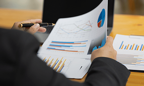 Person holding and reviewing printed charts and graphs with a pen, seated at a desk.