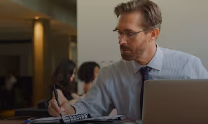 Man wearing glasses and a striped shirt writing in a notebook with a laptop in front of him in an office setting.