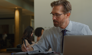 Man in glasses and striped shirt writing in a notebook next to a laptop, with blurred people in the background.
