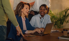 Two women sitting at a table working on a laptop, one pointing at the screen, with plants in the background.