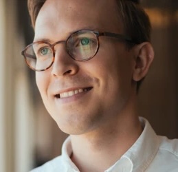 Smiling man with glasses looking outside near a window inside a warm indoor setting.