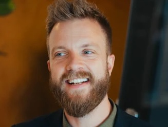Smiling man with brown hair, beard, and blue eyes wearing a dark jacket indoors.