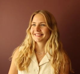 Smiling young woman with long blonde hair wearing a light-colored sleeveless top against brown backdrop.