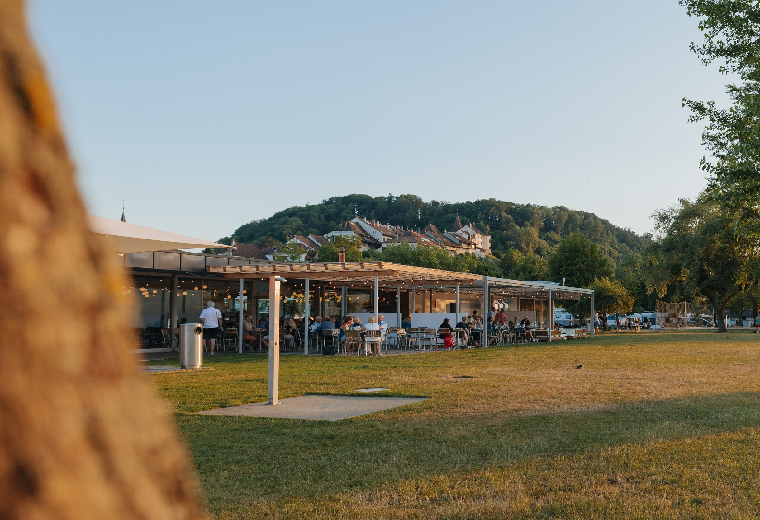 People sitting and dining under a long covered outdoor patio with string lights, beside a grassy open area and trees, with a hillside village in the background under a clear sky.