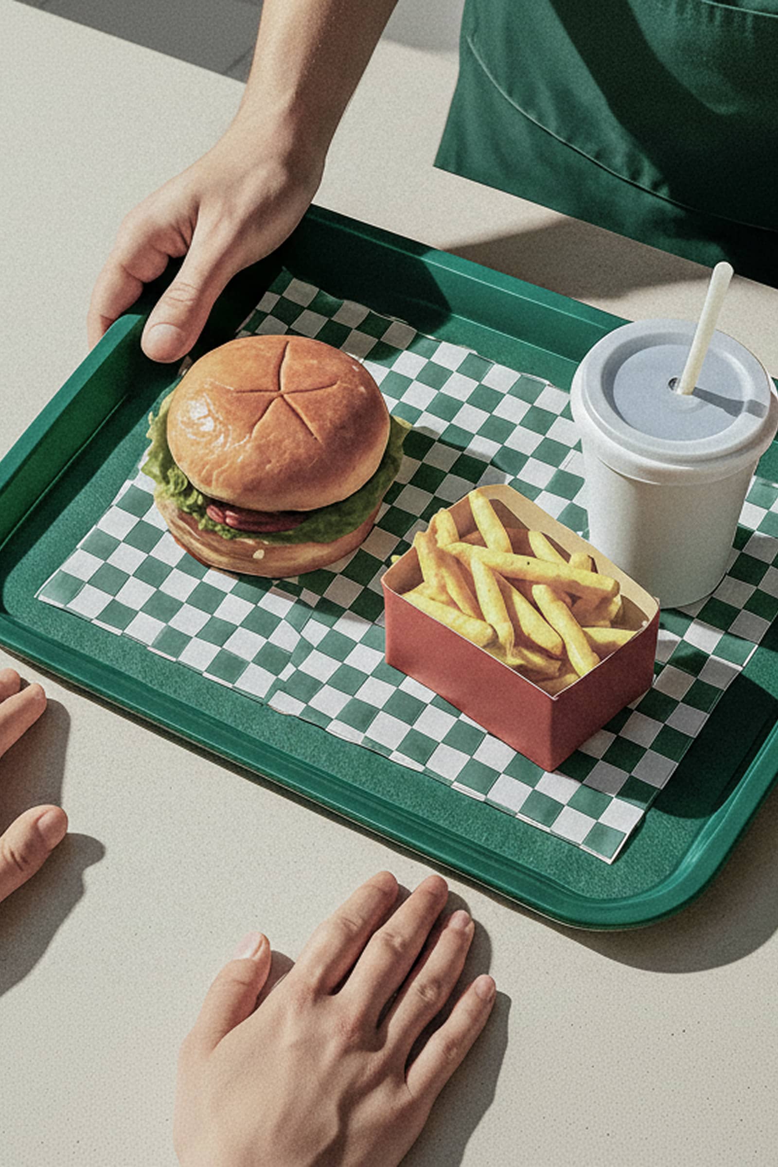 Tray with a burger, fries, and a drink being handed across a counter, with a hand reaching to receive it. The scene represents a casual meal pickup or quick service restaurant order.