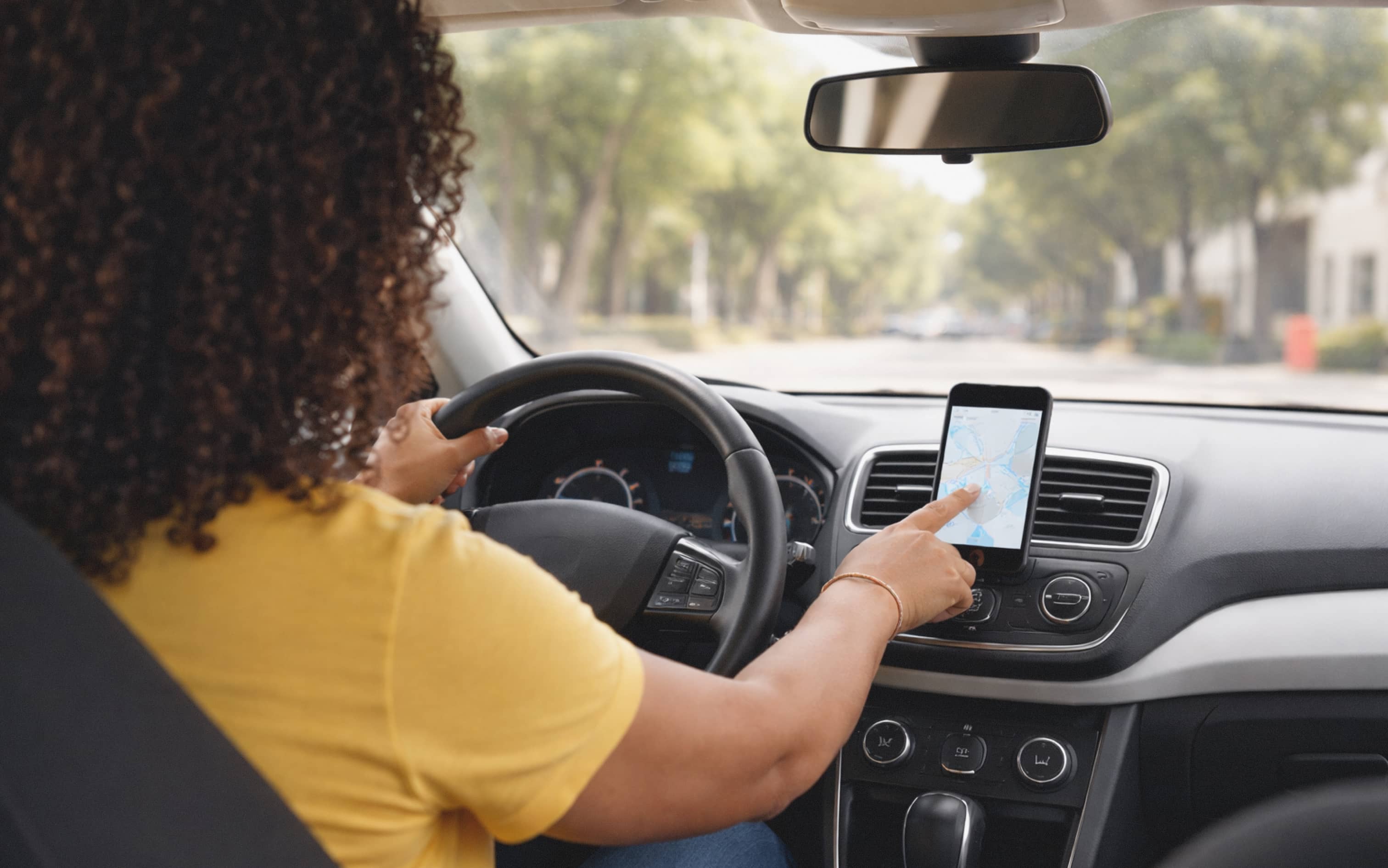 Person driving a car while pointing at a smartphone mounted on the dashboard displaying a map for navigation. The scene shows using directions while traveling on a tree lined street.