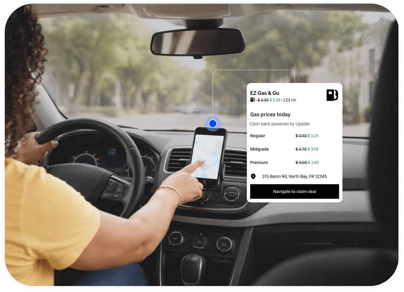 Person driving a car while pointing at a smartphone mounted on the dashboard displaying a map for navigation. The scene shows using directions while traveling on a tree lined street.