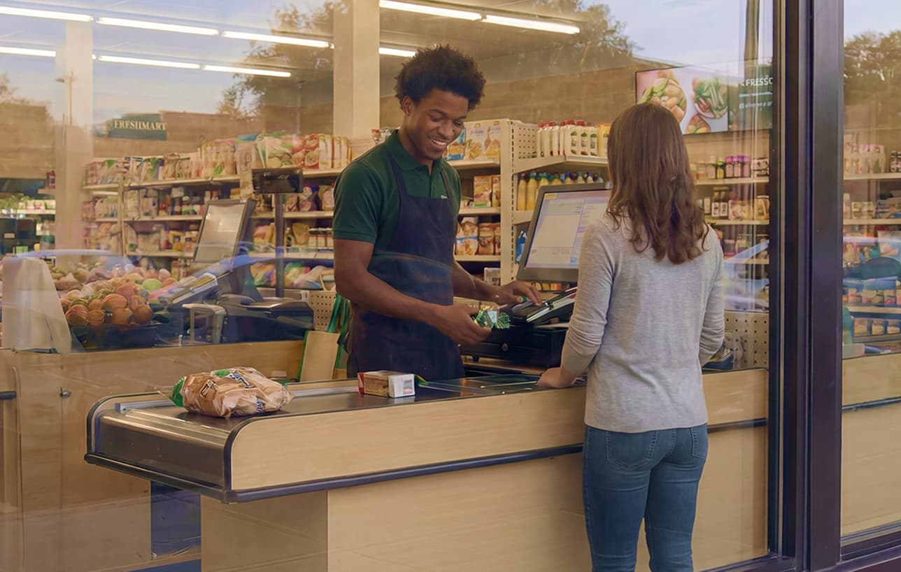 A grocery store cashier smiles while scanning items for a customer standing at the checkout counter. Shelves stocked with groceries and a produce display are visible in the background.