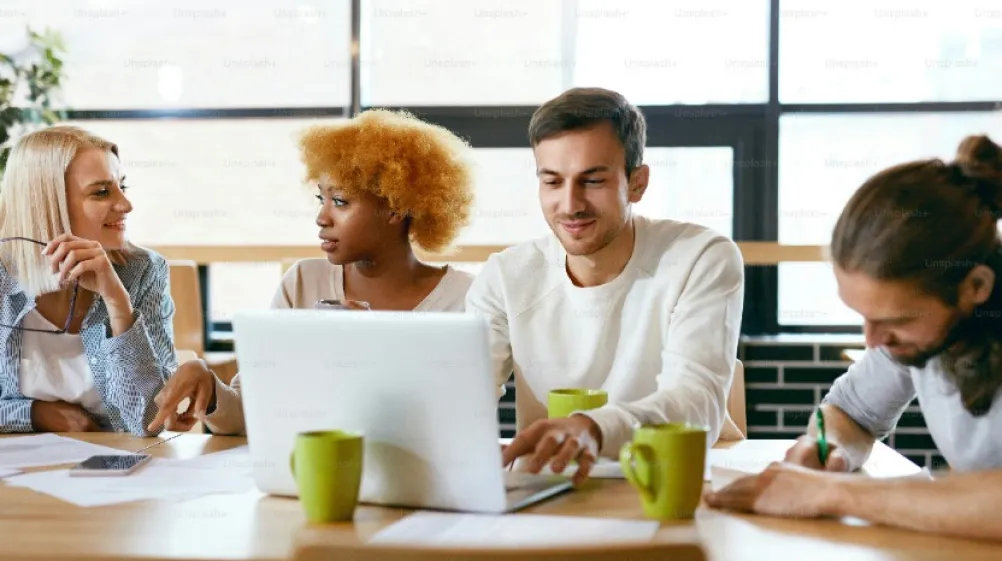 A group of young adults sat at a table chatting whilst using an laptop.
