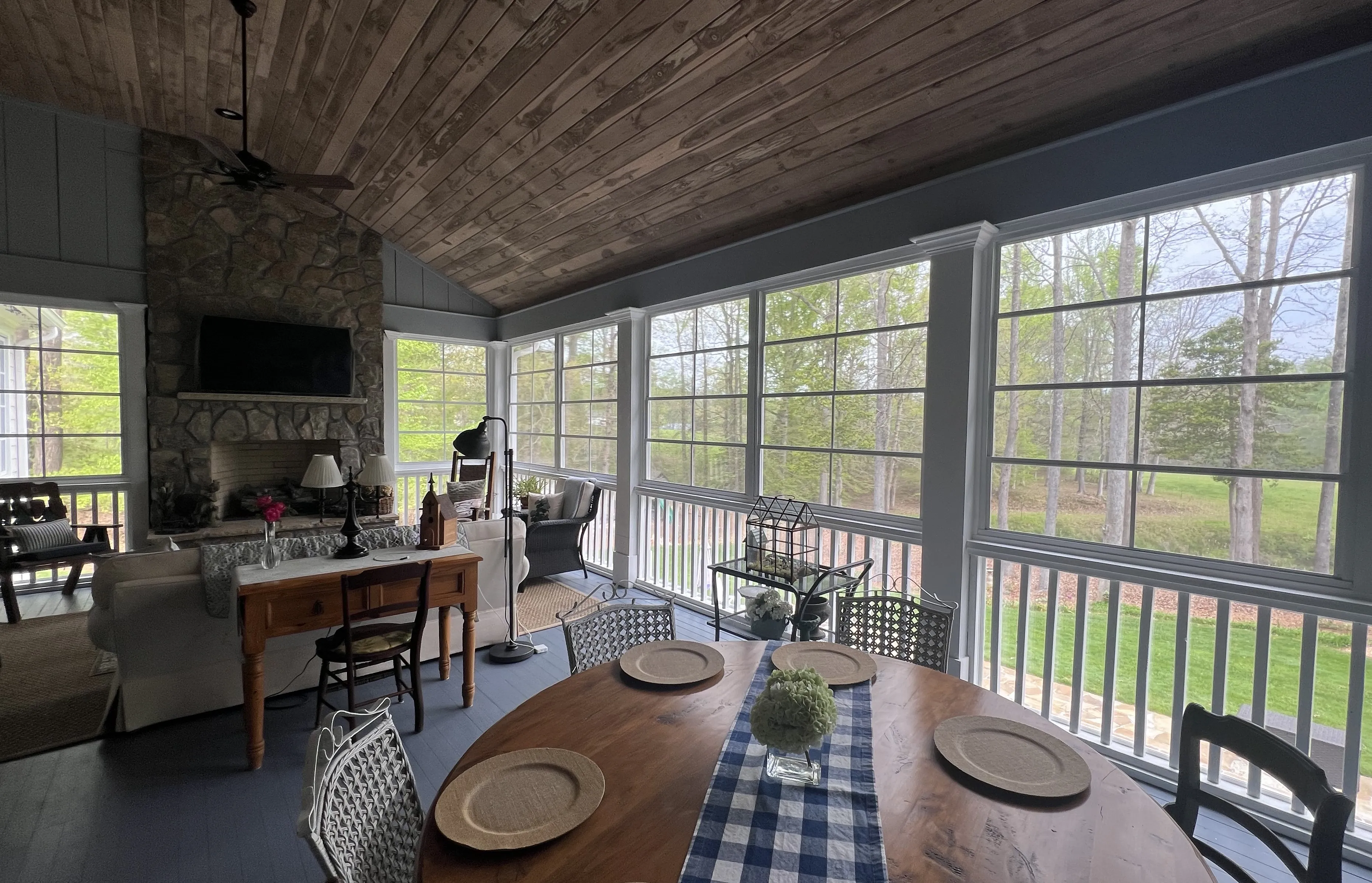 Bright screened porch with wooden ceiling, round dining table set with four plates and a centerpiece, stone fireplace, and seating area overlooking green trees and grass.