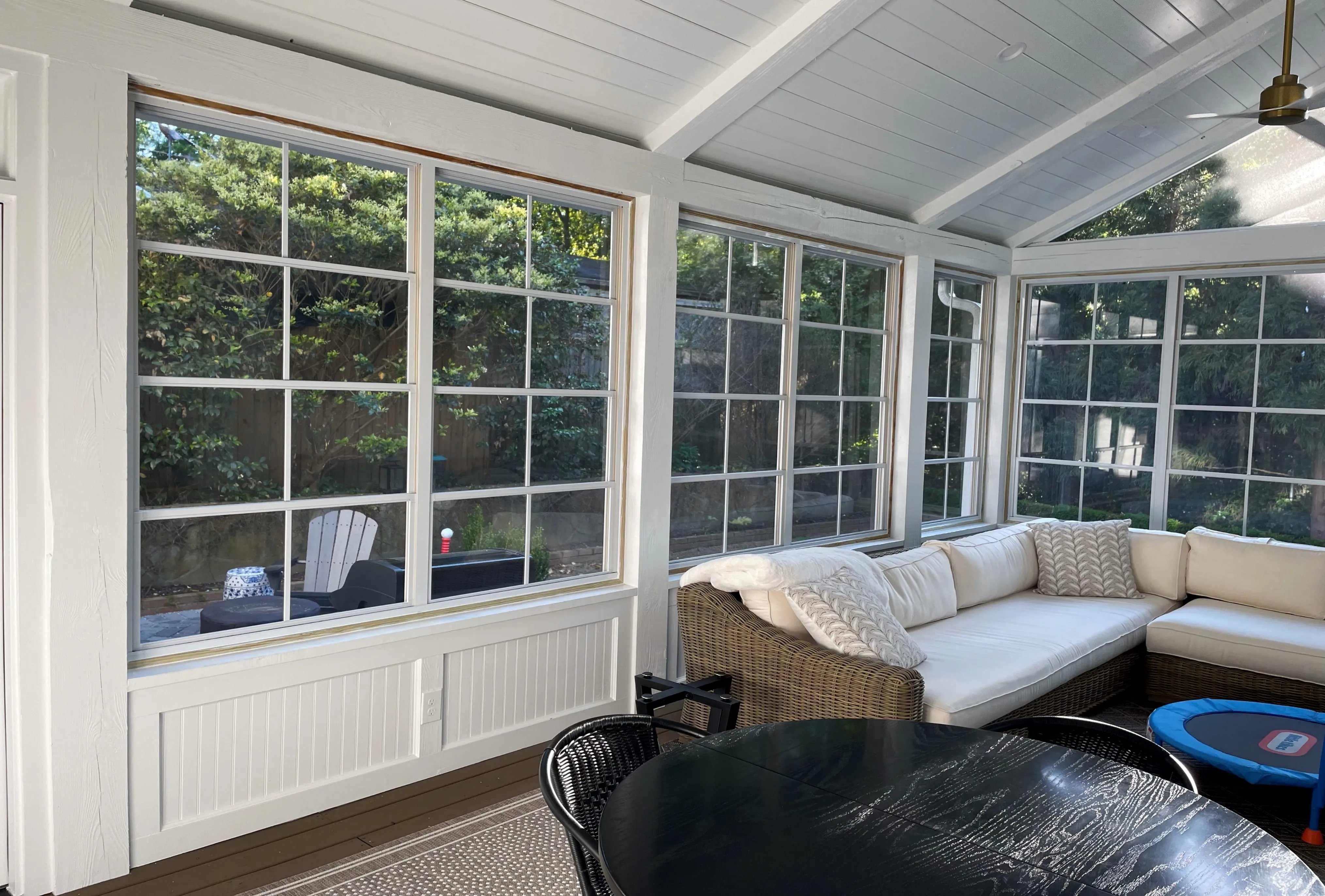 Bright sunroom with large grid windows showing garden outside, featuring a wicker sectional sofa with white cushions and a black round dining table.