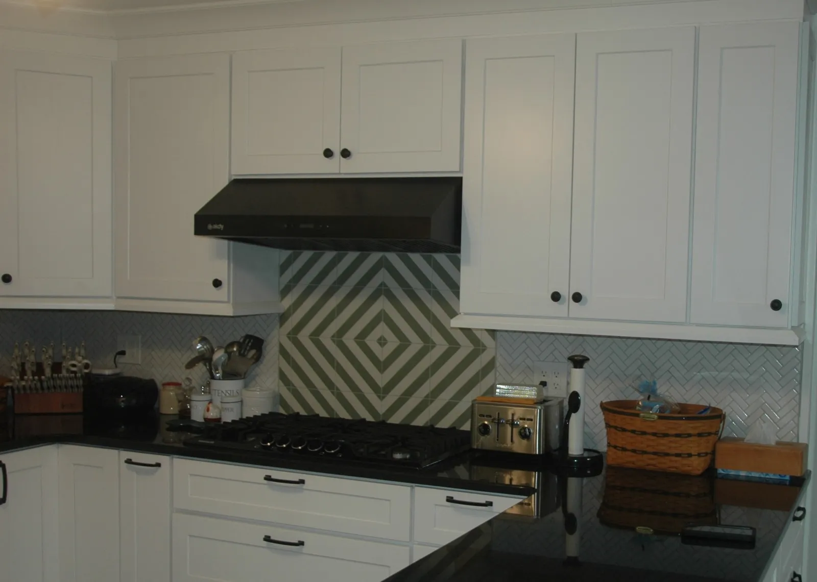 Modern kitchen corner with white cabinets, black countertop, stainless steel toaster, utensil holder, gas stove, black range hood, and decorative tile backsplash.