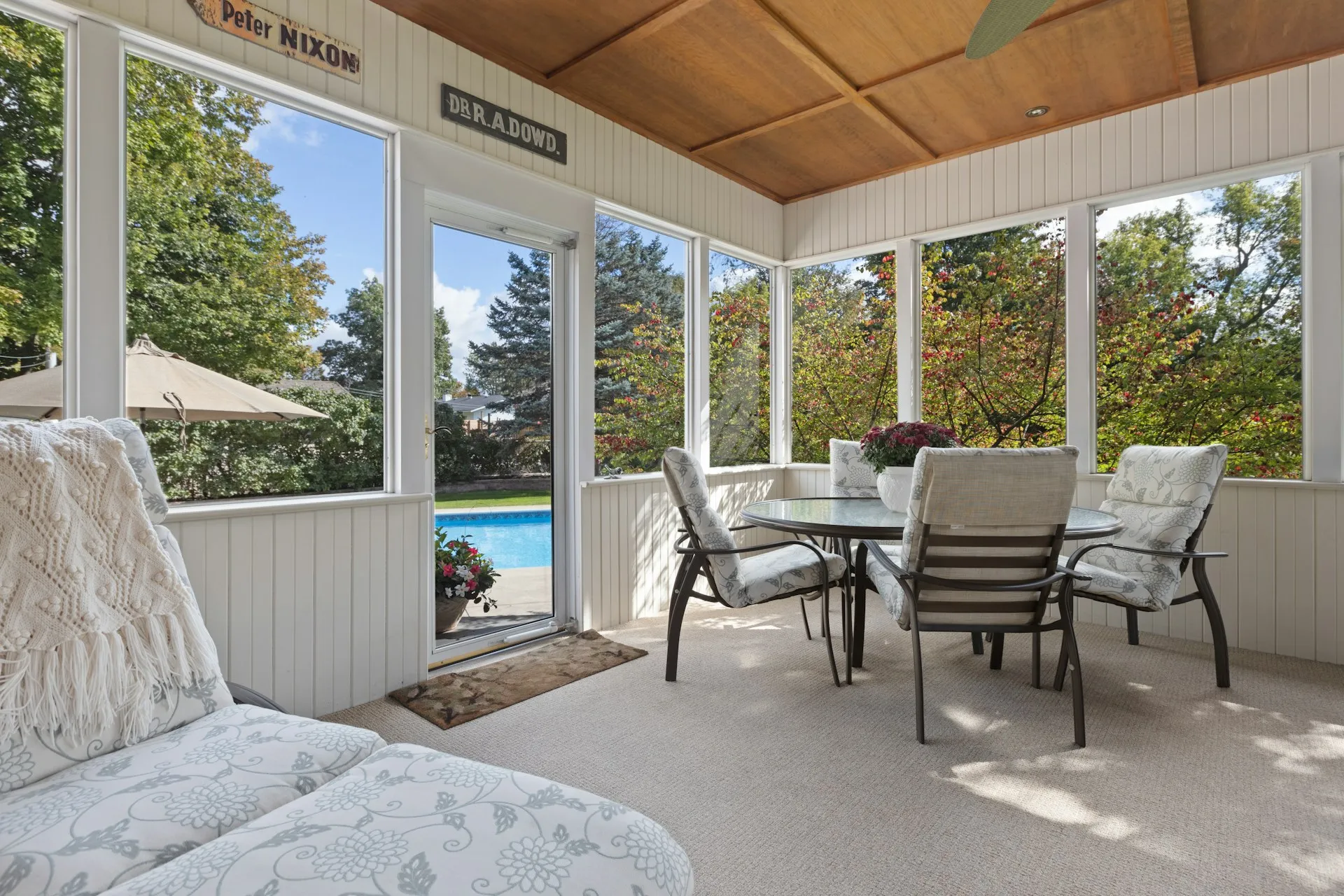 Bright covered patio with glass round table, cushioned chairs, and a view of a swimming pool and trees outside.