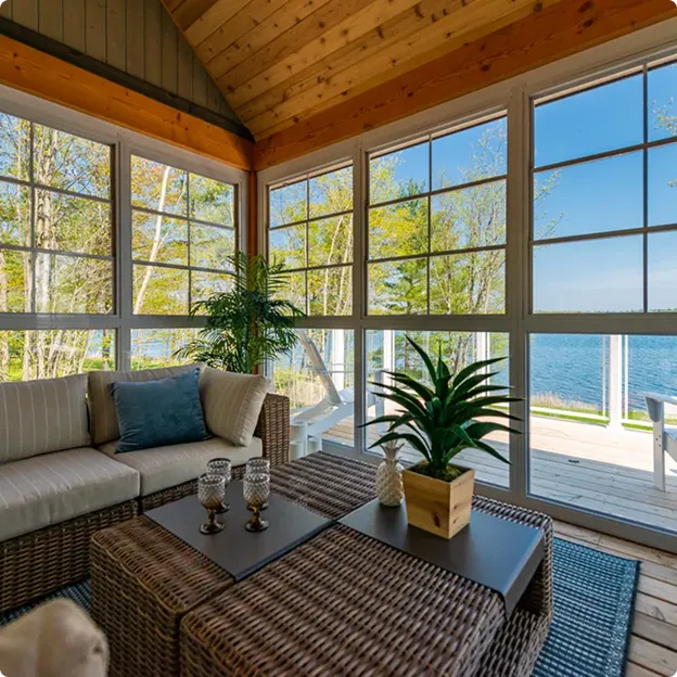 Cozy indoor seating area with wicker furniture and cushions overlooking a lake through large glass windows framed by wooden walls and ceiling.