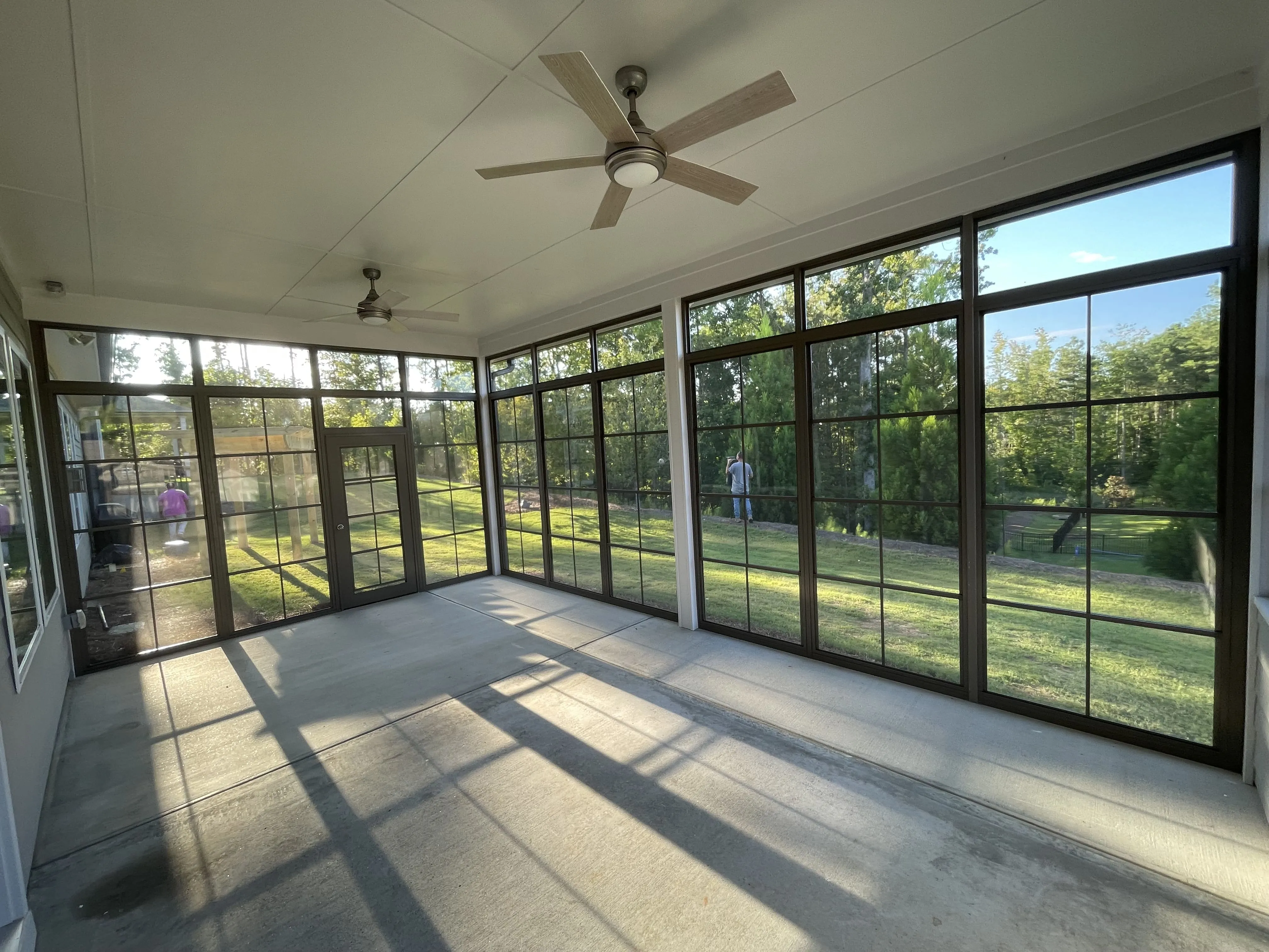 Empty screened-in porch with large windows, ceiling fans, and concrete floor overlooking a grassy yard and trees.