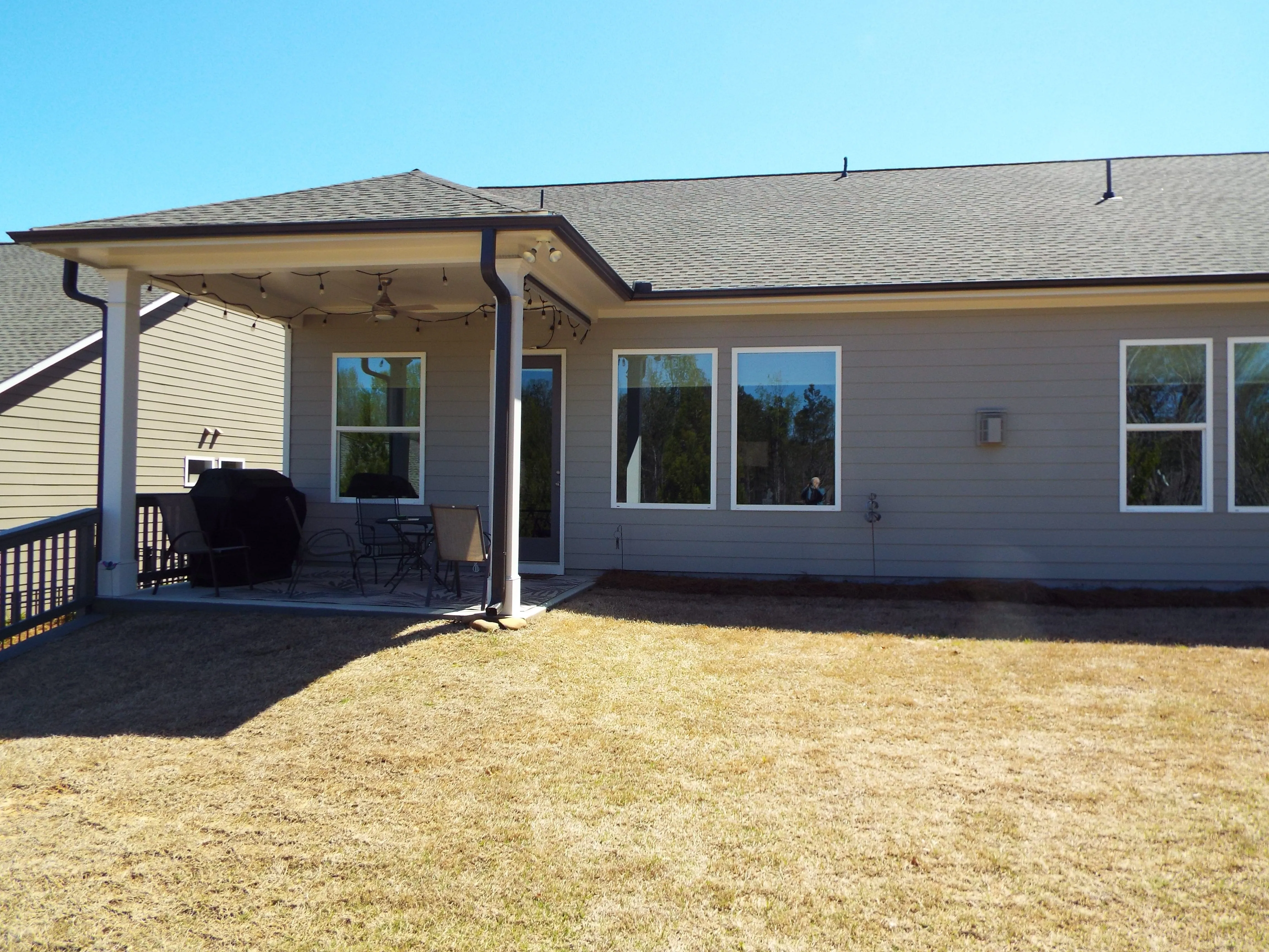 Backyard of a house with a covered patio featuring outdoor chairs, a table, a grill, and string lights under a clear blue sky.