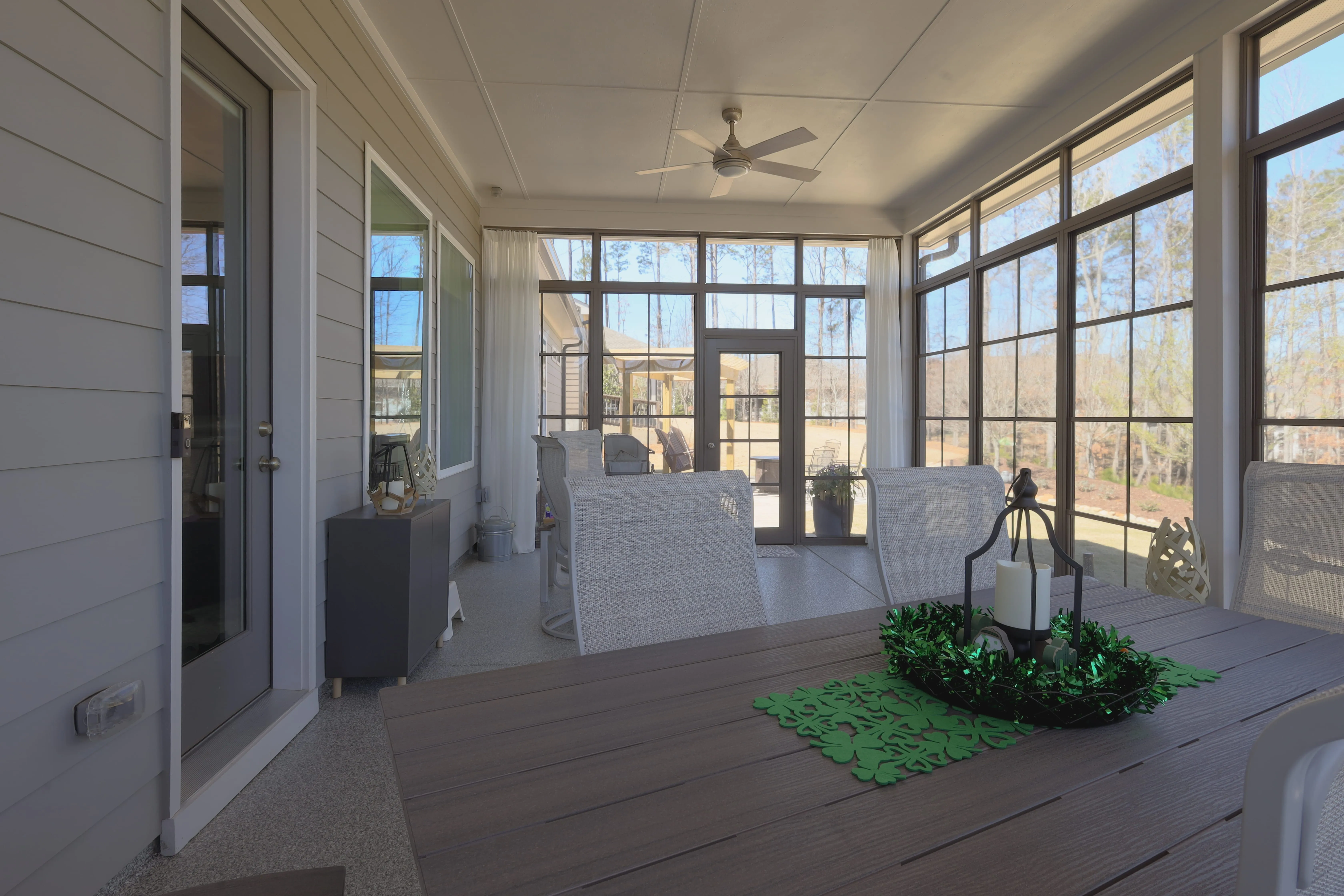 Bright enclosed porch with large windows, a ceiling fan, and a wooden table decorated with a candle centerpiece and green runner.