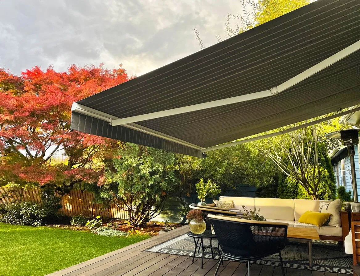 Outdoor patio seating area with beige couch, black chair, wooden deck, and large retractable awning overhead, surrounded by green and red-leaved trees.