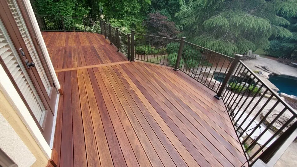 Spacious wooden deck with dark metal railing overlooking a backyard with trees and a pool.