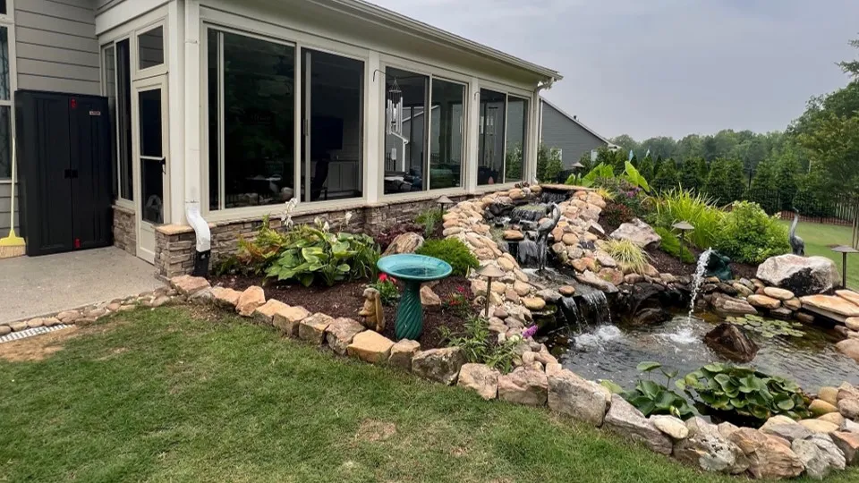 Sunroom with large windows overlooks a landscaped garden featuring a stone-bordered pond with cascading waterfalls and various plants.