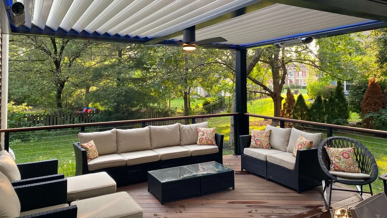 Outdoor patio with beige cushioned black wicker sofas and chair under a modern pergola with a ceiling fan and ambient lighting, overlooking a green garden.