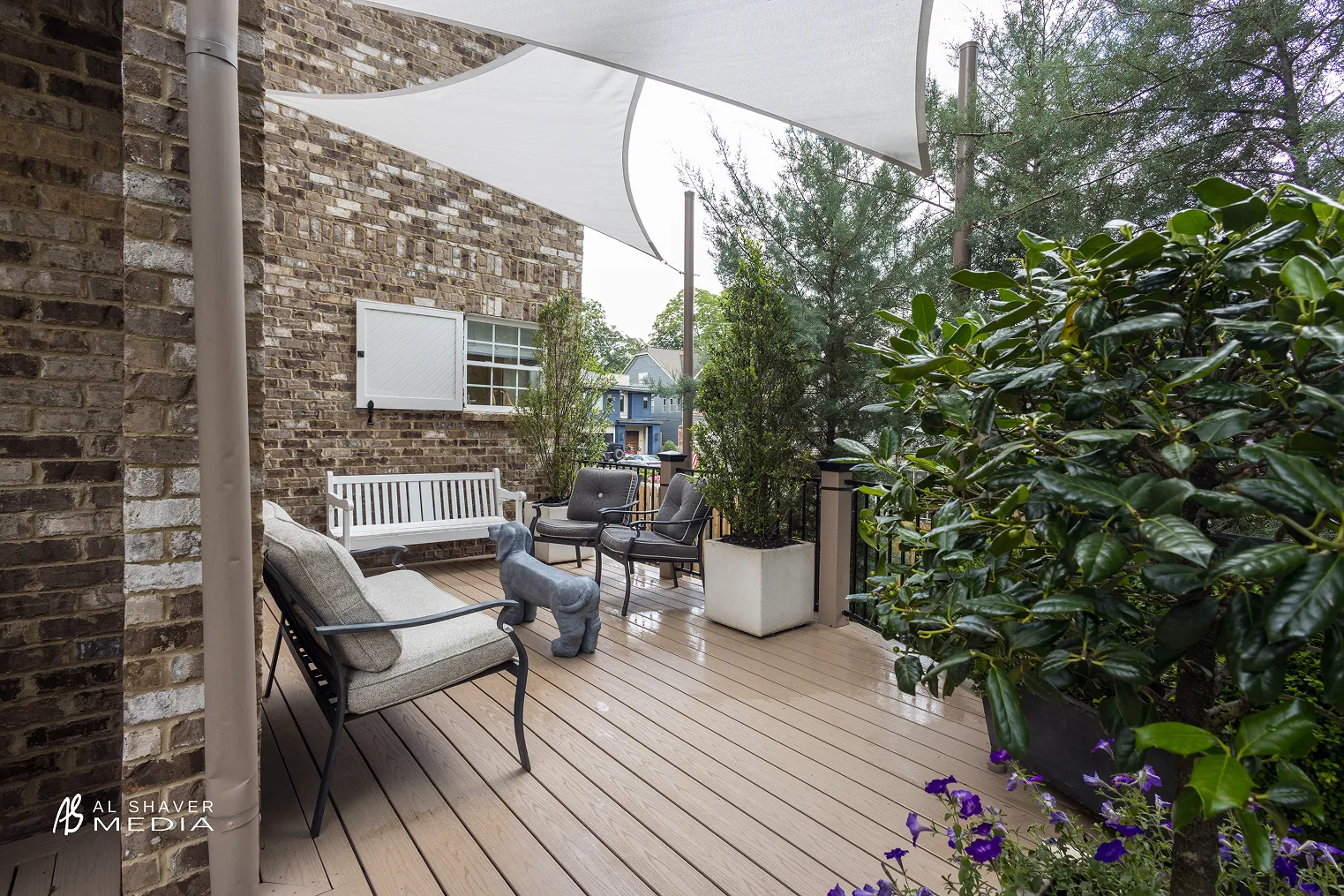 Cozy outdoor deck with cushioned chairs, a white bench, potted plants, a dog statue, and triangular shade sails overhead.