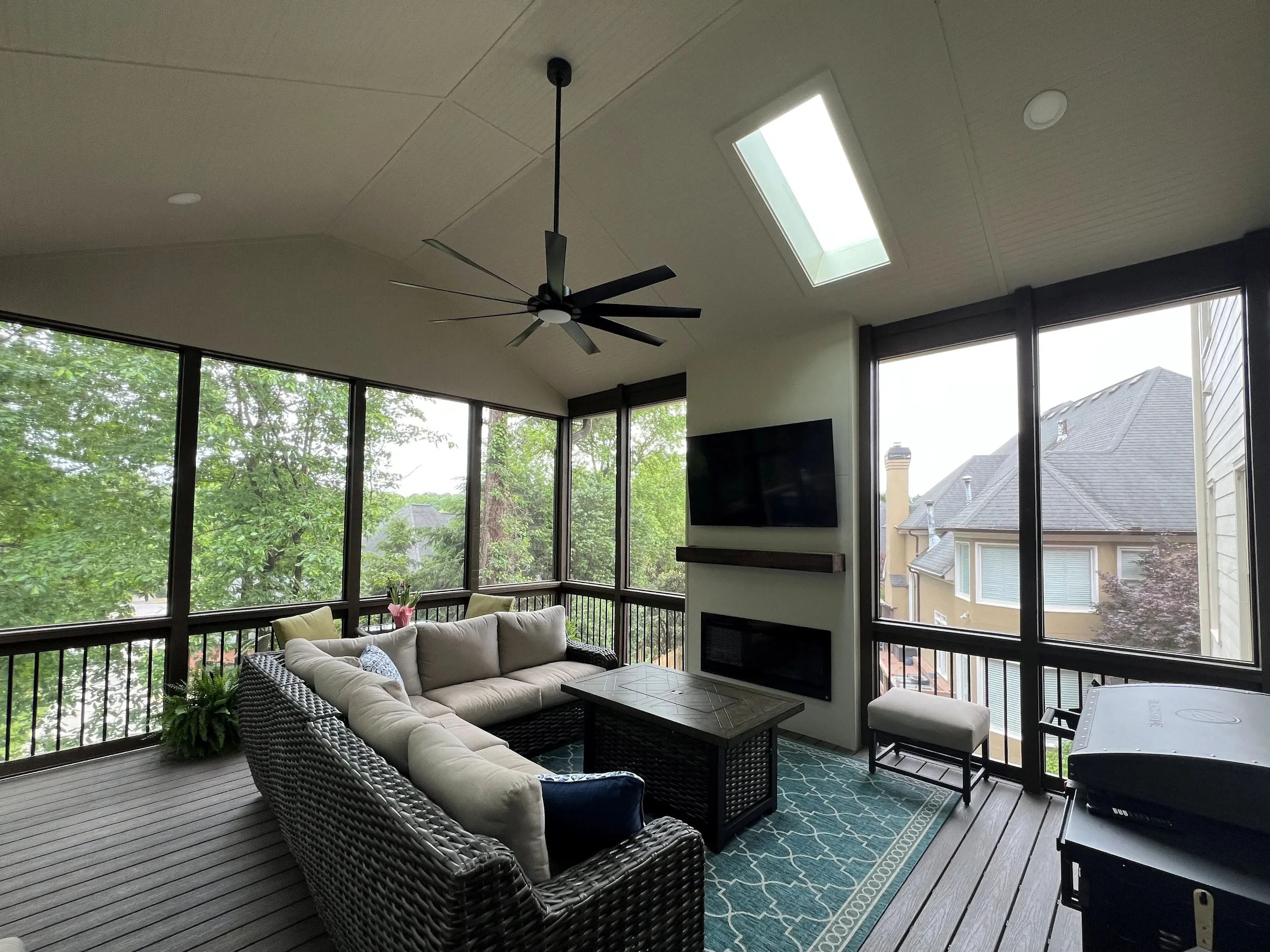 Screened porch with a wicker sectional sofa, coffee table, ceiling fan, mounted TV, fireplace, skylight, and view of trees and neighboring houses.