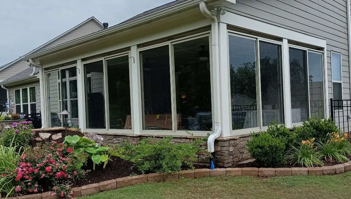 Sunroom with large windows and stone base attached to house, surrounded by garden beds with flowers and shrubs.