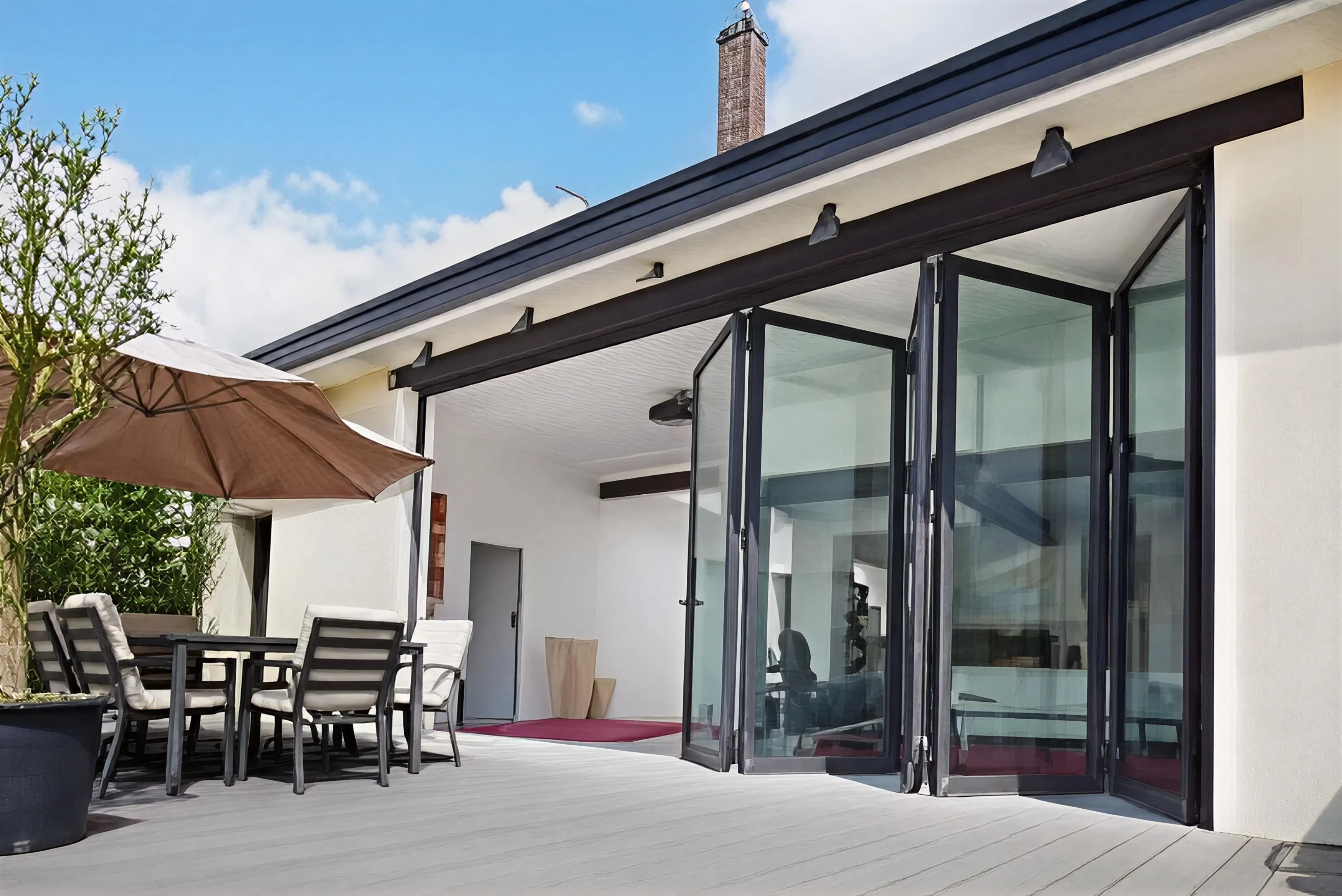 Modern patio with cushioned chairs around a table under a large beige umbrella, next to folding glass doors leading inside.