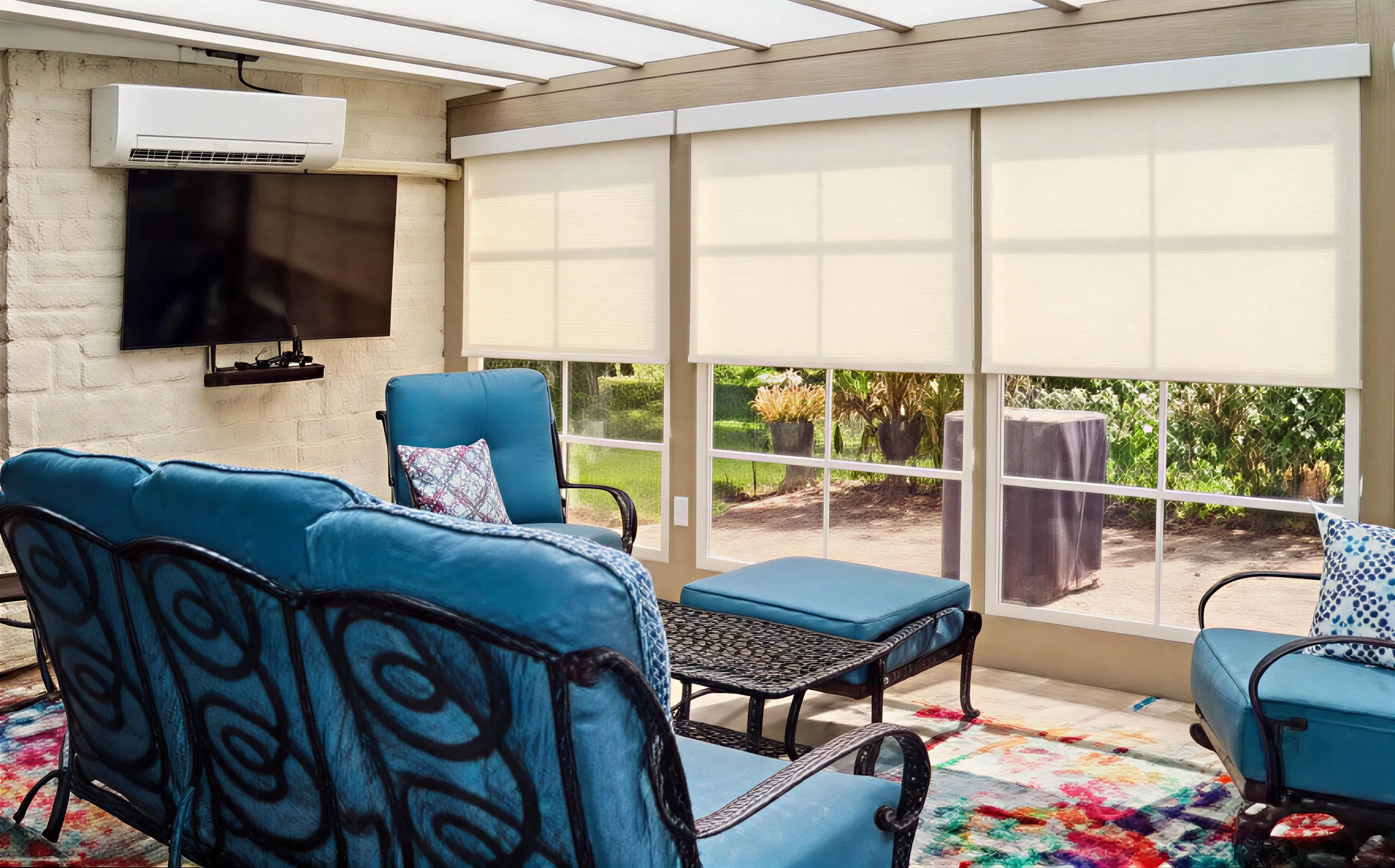 Sunroom with blue cushioned metal furniture, a colorful rug, large windows with white roller shades, a wall-mounted TV, and a white air conditioner unit.