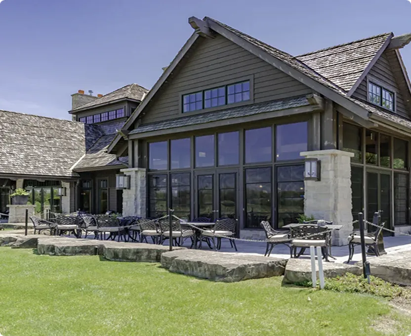 Modern building with large glass windows, stone pillars, and an outdoor patio furnished with black metal tables and chairs on a green lawn under a clear sky.