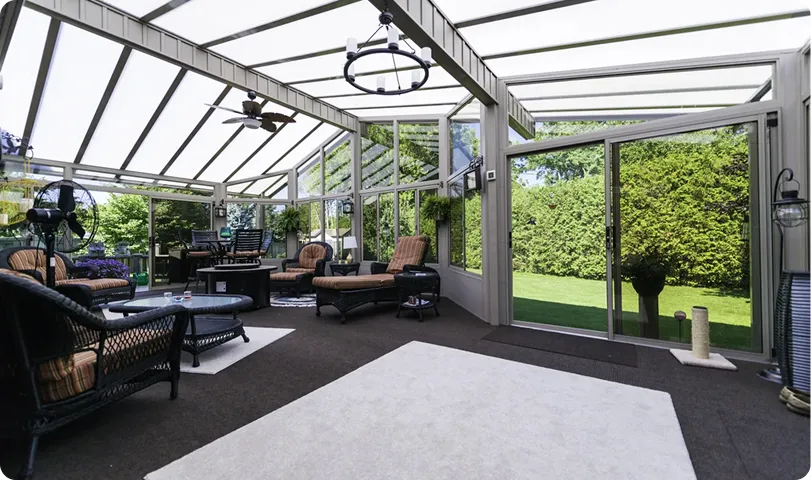 Sunroom with wicker furniture, ceiling fan, glass walls and ceiling, overlooking a green lawn and garden.