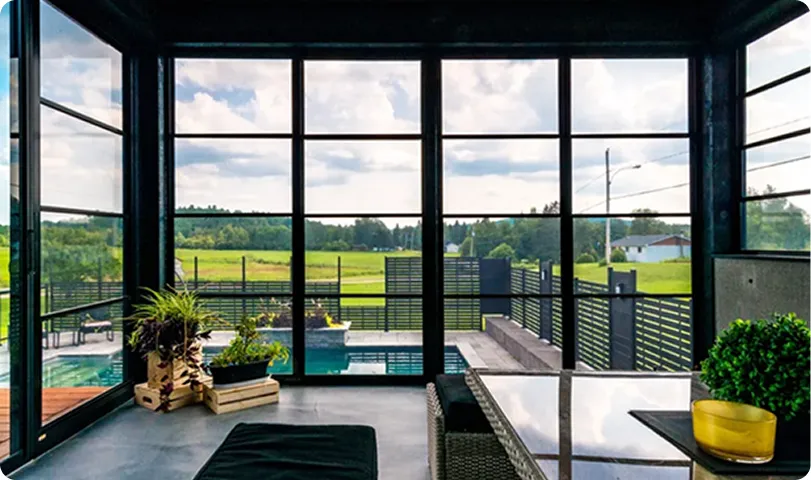 Modern patio with large glass windows overlooking a green field and a swimming pool, featuring plants and outdoor furniture.